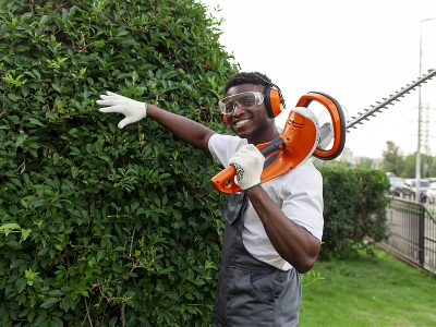 garden worker in uniform cuts bushes, african american man in goggles and headphones works in the garden with garden electric tool, pruning trees and bushes